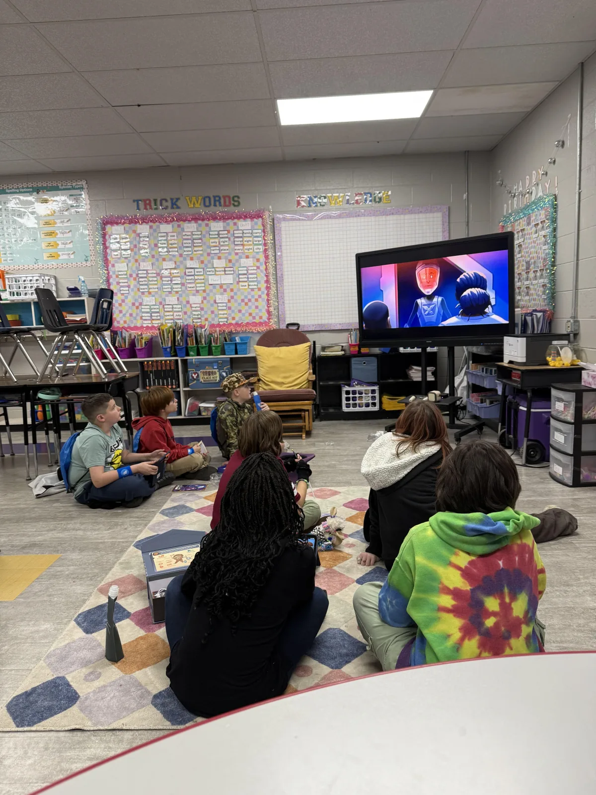 Students sit together watching a story on the classroom screen